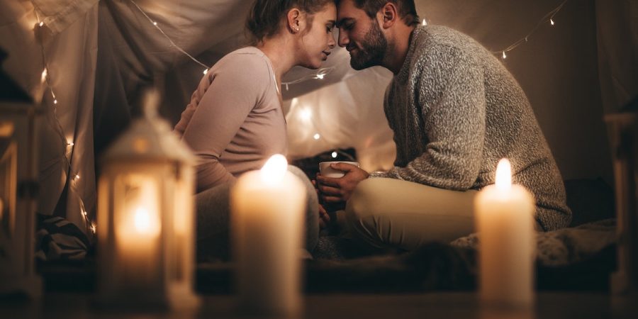 Dating your fiancé during your wedding planning period. Romantic couple sitting on bed facing each other touching their foreheads and with eyes closed. Man sitting on bed with his wife holding a coffee cup in a room decorated with candles and tiny serial bulbs.
